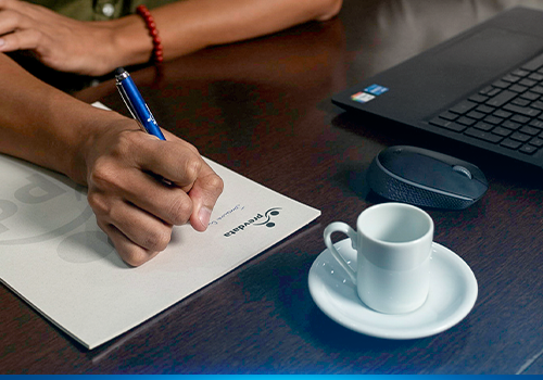 Detalhes de uma mesa de trabalho com computador, xícara de café e pessoas trabalhando
